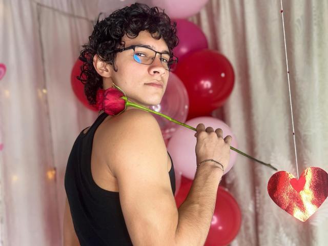 SamuelHills, met krullend zwart haar, een bril en een zwart tanktop, houdt een rode roos voor zijn nek, en staat voor roze en rode ballonnen..