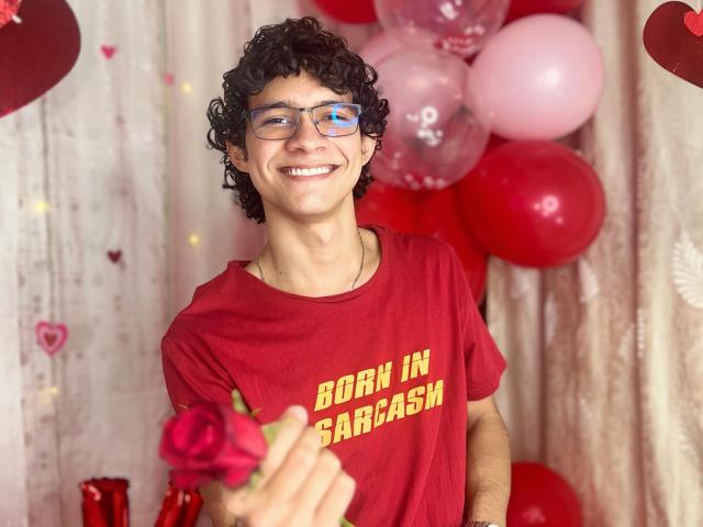 SamuelHills, met krullend zwart haar, een bril en een rood shirt, houdt een roze roos vast en glimlacht naar de camera, omringd door rode en roze ballonnen. Zijn achterkant is niet zichtbaar..
