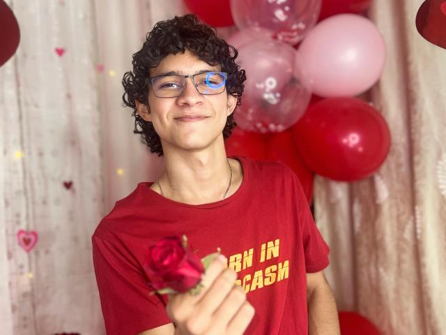 SamuelHills met krullend zwart haar, een blauwe bril, een rood shirt, die een roos vasthoudt, lachend, met op de achtergrond rode en roze ballonnen, een wit gordijn met hartdecoraties..