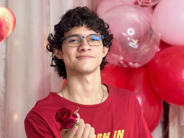 SamuelHills, met krullend zwart haar, een blauwe bril, een rood shirt, die een rode roos vasthoudt, met een lichte glimlach, omringd door roze en rode ballonnen, ziet er schattig en een beetje nerdy uit..