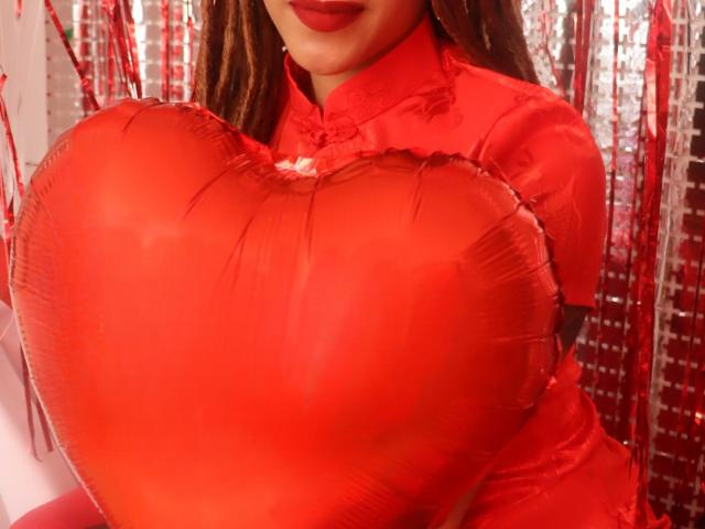 KendallQuinn holding a shiny red heart-shaped balloon, wearing a red shirt, her lips painted red, her brown hair partially visible, the balloon covering her large, shiny tits.
