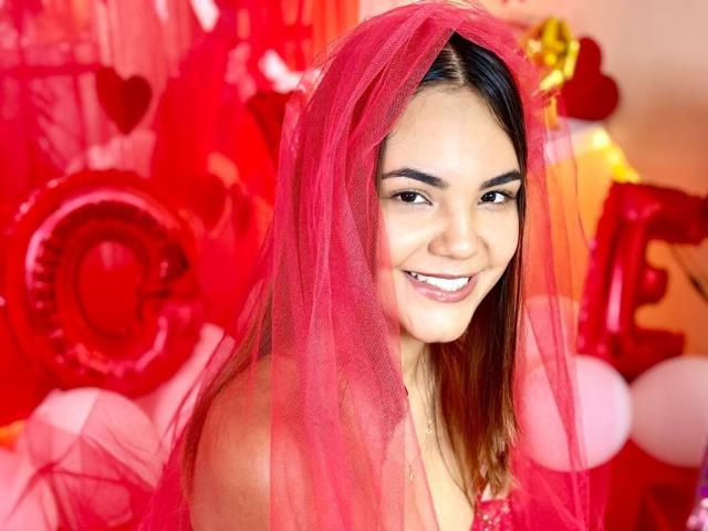 StaceyWhite with pink veil, smiling, brown eyes, straight dark brown hair, wearing red dress, red balloons and heart-shaped decorations in the background. She has cute tits and a sexy face.