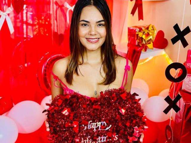 StaceyWhite with long brown hair, smiling, holding a red heart-shaped boob decoration, pink straps, red and white balloons, and "XOXO" in black. Red and pink background.