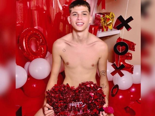 JassonSmith, shirtless, with a small heart tattoo on his chest, holding red tinsel, smiling, short brown hair, tattoos on his right arm, in a red and white decorated room.