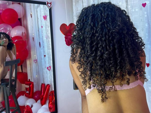 RebeccaMils with curly black hair, back to the mirror, wearing pink lace bra, reflecting in mirror, surrounded by red and white balloons, heart decorations, and a rose.