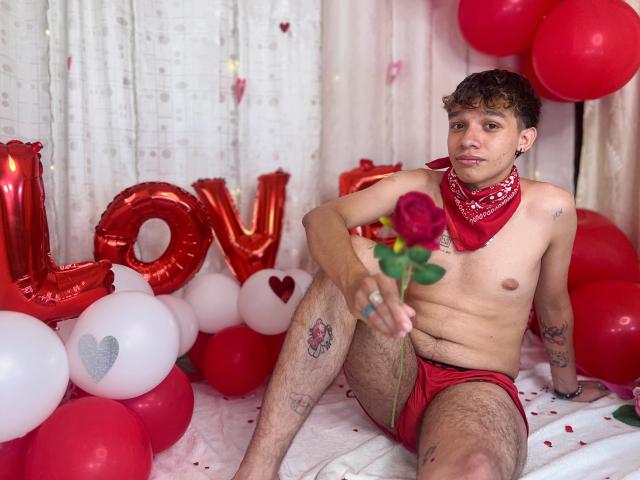 KristofMills, shirtless, red underwear, holding rose, red bandana, tattoos, curly brown hair, white skin, sitting on bed, surrounded by red and white balloons, looking at camera.