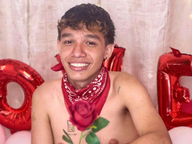 Shirtless KristofMills with curly brown hair, red bandana, holding rose, smiling, red balloons, and red number balloons in background. Tattoo on right chest. Slightly tanned, cute.