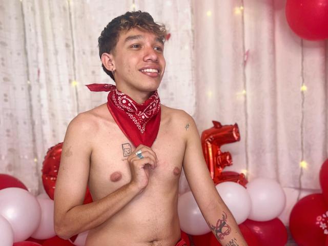 KristofMills, shirtless with a red bandana, smiling, holding his chest, tattoos on chest and arm, curly brown hair, fair skin, surrounded by red and white balloons.