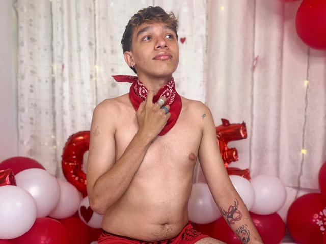 Shirtless KristofMills with curly brown hair, wearing a red bandana, sitting in red and white balloons, looking up with a slight smile, tattoo on right arm, medium-sized flaccid penis.