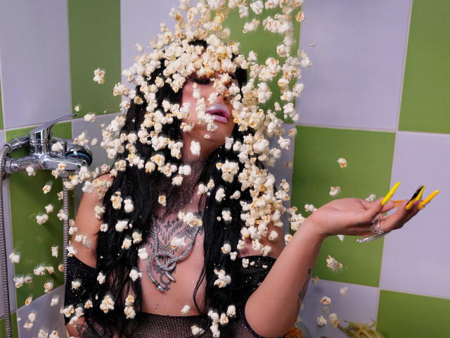 DenyChantal with black hair, yellow nails, and large chest, covered in flying popcorn, wearing a black mesh top and a giant silver necklace, in a green and white tiled bathroom.