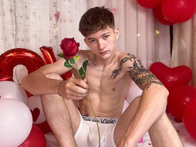 Shirtless JakeDavins with tattoos, holding a red rose, wearing white Calvin Klein underwear, sitting amidst red and white balloons, looking serious. JakeDavins has a slim build, brown hair.