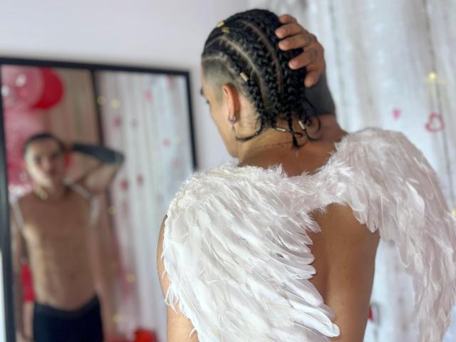 AlexBernal with braided hair, wearing white angel wings, facing mirror, reflection shows shirtless upper body, hand on head, white curtain, red balloon in background.