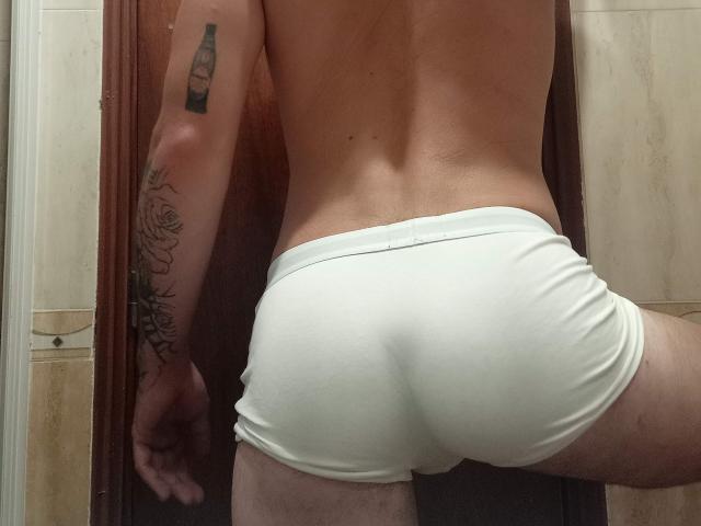 AlessRicalle's white boxer briefs, showing their ass, with a bottle tattoo on the left arm and a rose tattoo on the right arm. Bare back, standing in a room.
