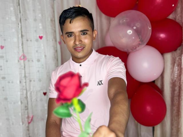 DayanWilly tenant une rose rouge, portant une chemise rose, avec des cheveux noirs courts, un léger sourire, tendant la main, des ballons rouges et roses en arrière-plan, un rideau avec des motifs en forme de cœur sur la gauche..