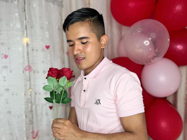 DayanWilly, met kort zwart haar, draagt een roze polo, houdt een rode roos vast en staat voor roze en rode ballonnen. Ze glimlacht zachtjes en kijkt naar de roos..