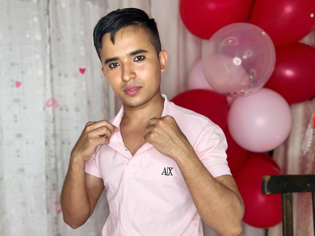 DayanWilly, a cute Asian dude with short black hair, wearing a pink shirt, stands with hands near his chest, looking at the camera with a slight pout. Red and pink balloons.