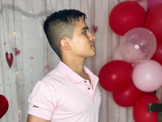 DayanWilly with short dark hair, light pink polo, looking right, standing by red and pink balloons, soft light, slight smile, background blurry.