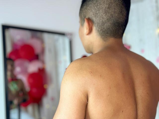 DayanWilly's kale rug, met kort, donker haar, kijkt naar een spiegel die een vage weerspiegeling toont van rode en roze bloemen. DayanWilly's huid is glad met een paar kleine littekens..