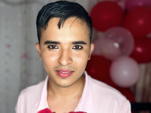DayanWilly ha i capelli neri e corti, gli occhi marroni e un leggero baffo, indossa una camicia rosa con una cravatta rossa e guarda la telecamera con un leggero sorriso..