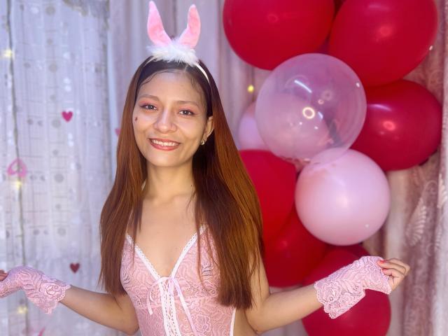 KarolCrystal has brown hair, pink lace lingerie, white bunny ears, and pink wrist cuffs, smiling with small tits, and a cute ass, standing with red and pink balloons.