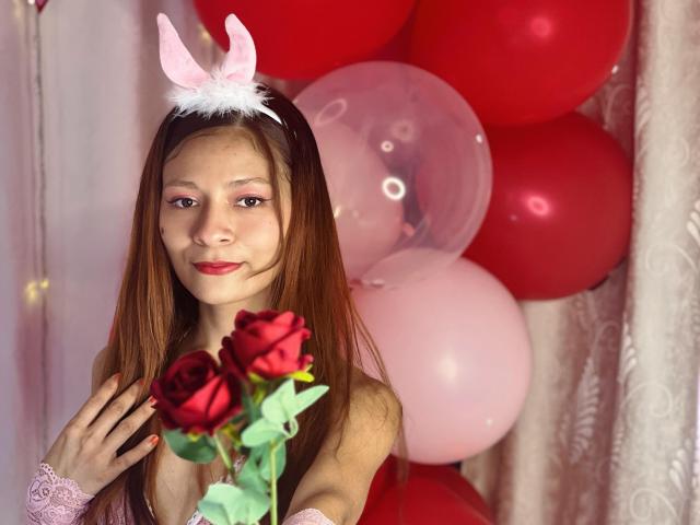 KarolCrystal with long brown hair, pink bunny ears, holding red roses, wearing pink lace cuffs, standing in front of red and pink balloons, smiling slightly.