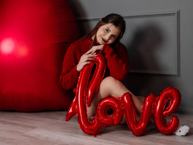 AdeliaLove with red balloon letters spelling "love" on wooden floor, wearing red sweater, sitting with knee up, smiling, dark brown hair, gray wall, red beanbag on left.