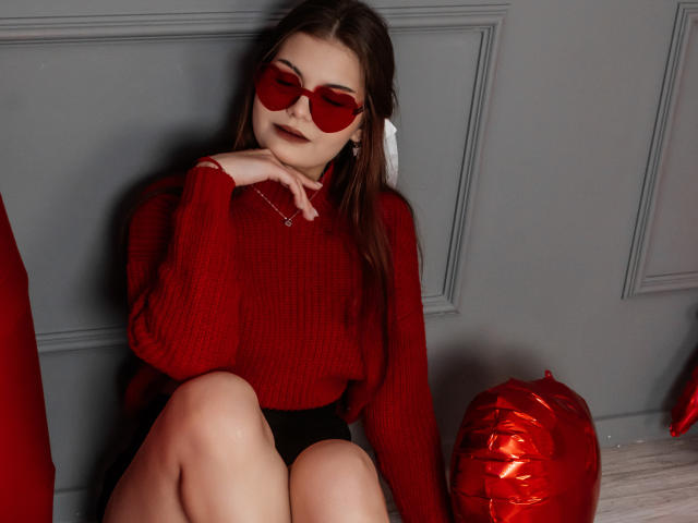 AdeliaLove with red sunglasses, red sweater, black shorts, and a white hair ribbon, sitting with her hand near her face, and red balloons on the floor.