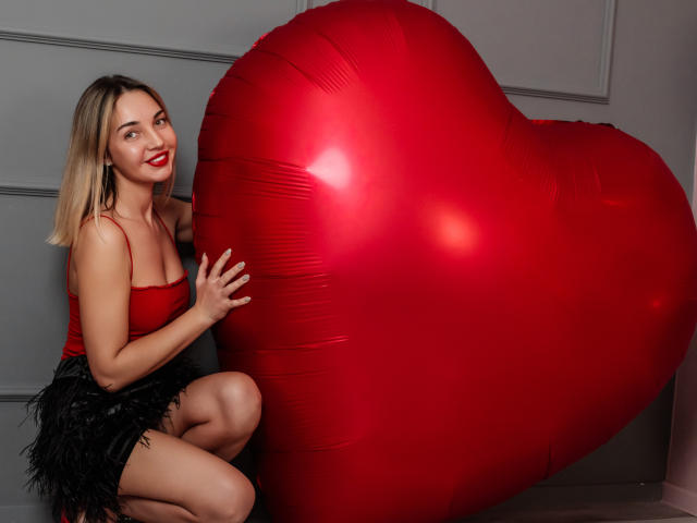LettieSweet with blonde hair, red top, black feather skirt, kneeling, smiling, holding a huge red heart-shaped balloon, red lips, fair skin, cute, sexy.