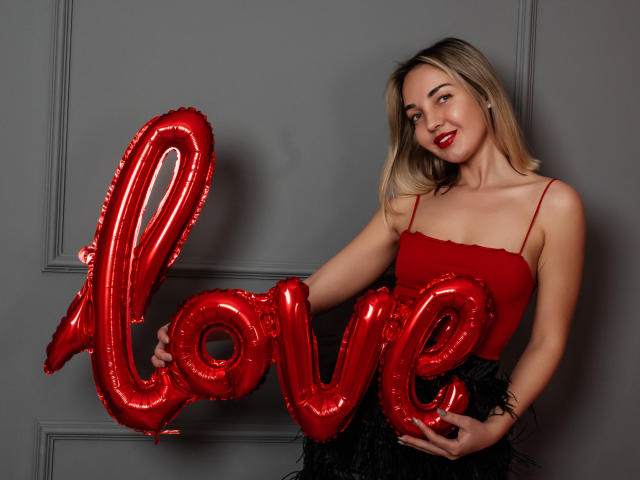 LettieSweet with red shiny balloons shaped like "love", wearing a red spaghetti strap top and black skirt, smiling with red lipstick, blonde hair, holding balloons against gray wall.