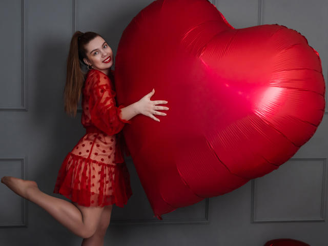 MilanaKitty con un vestito a pois rossi, che tiene un palloncino a forma di cuore rosso gigante, in piedi su una gamba, sorridente, con rossetto rosso, con i capelli raccolti in una coda di cavallo, sfondo di un muro grigio, un cuore rosso a sinistra..