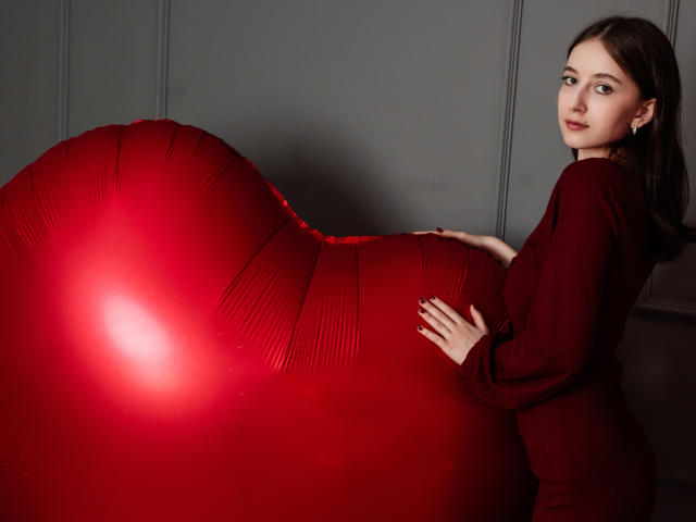 LilieFlower, avec une énorme bosse rouge, portant une robe rouge foncé, a sa main posée dessus et regarde par-dessus son épaule avec une expression calme. Elle porte des boucles d'oreilles..