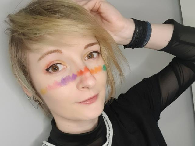 AmazingBlair with short blonde hair, wearing a black sheer top, has colorful paint on her cheek, brown eyes, and a slight smile, with a black bracelet on her wrist.