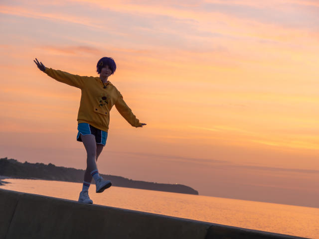 AmazingBlair with a purple helmet, yellow hoodie, blue shorts, and white socks, balancing on a concrete edge at sunset, arms out, looking forward, with a calm ocean behind.