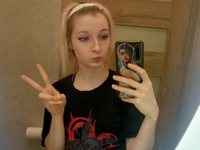 AmazingBlair with pink eyeliner, blonde hair, white headband, black graphic tee, making a peace sign, holding a phone with a face cover, in a bathroom selfie.