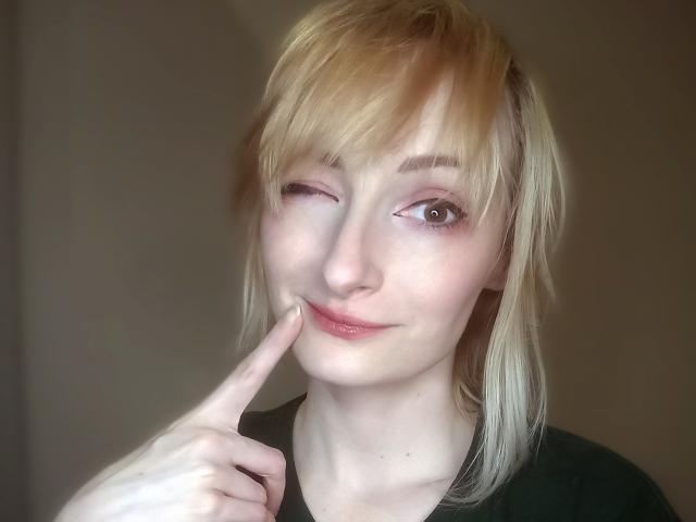 AmazingBlair with blonde hair, winking, and pointing to her slightly parted lips with a finger. She's wearing a green shirt, and her skin is pale.