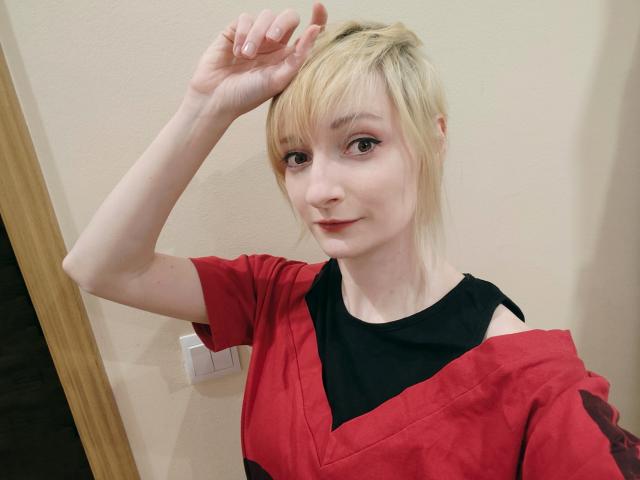 AmazingBlair with blonde hair, light skin, and red shirt, black top, smiling, right hand touching head, brown eyes, subtle makeup, standing against beige wall, small white light switch.