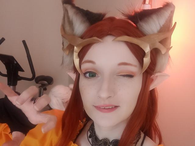 AmazingBlair with red hair, cat ears, and gold headpiece, winking, has pale skin, green eyes, and a black lace choker, showing a peace sign with her right hand.