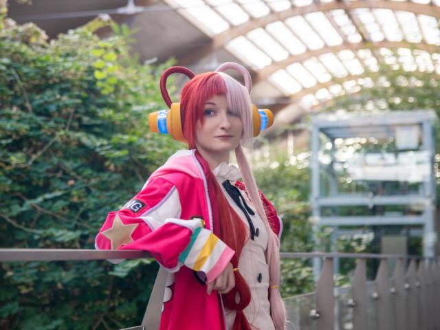 AmazingBlair with pink and red hair, wearing colorful headphones, a pink jacket, and white shirt, holding a star, standing outside with greenery and a metal structure in background.