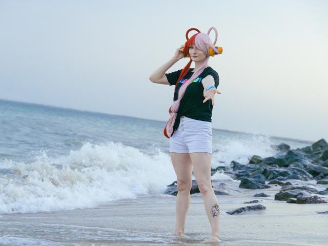 AmazingBlair with pink hair, orange headband, black shirt, white shorts, and pink bag, standing barefoot on beach, waving hand, showing tattoo on right thigh, ocean in background.