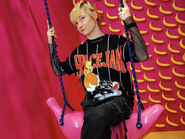 AmazingBlair with blonde hair, wearing a black "DICE JAM" shirt, black mesh sleeves, and black pants, sits on a pink swing with blue ropes, looking cute.
