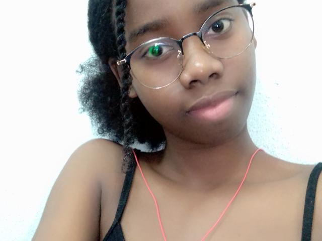 Lindah69 with dark skin, curly black hair in braids, wearing glasses and a black spaghetti strap top, pink earbuds, and a slight smile, looking at the camera.