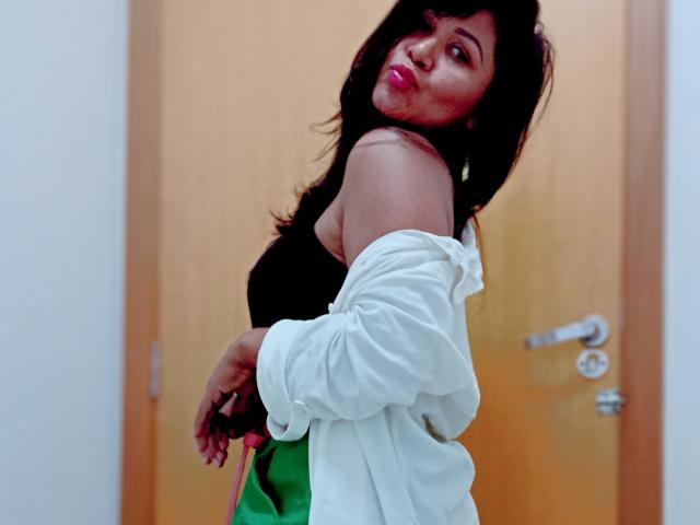 Lafertte with dark hair, pouty red lips, off-shoulder white shirt, black top, green and white skirt, looking over shoulder, brown door in background. Boobs slightly visible.