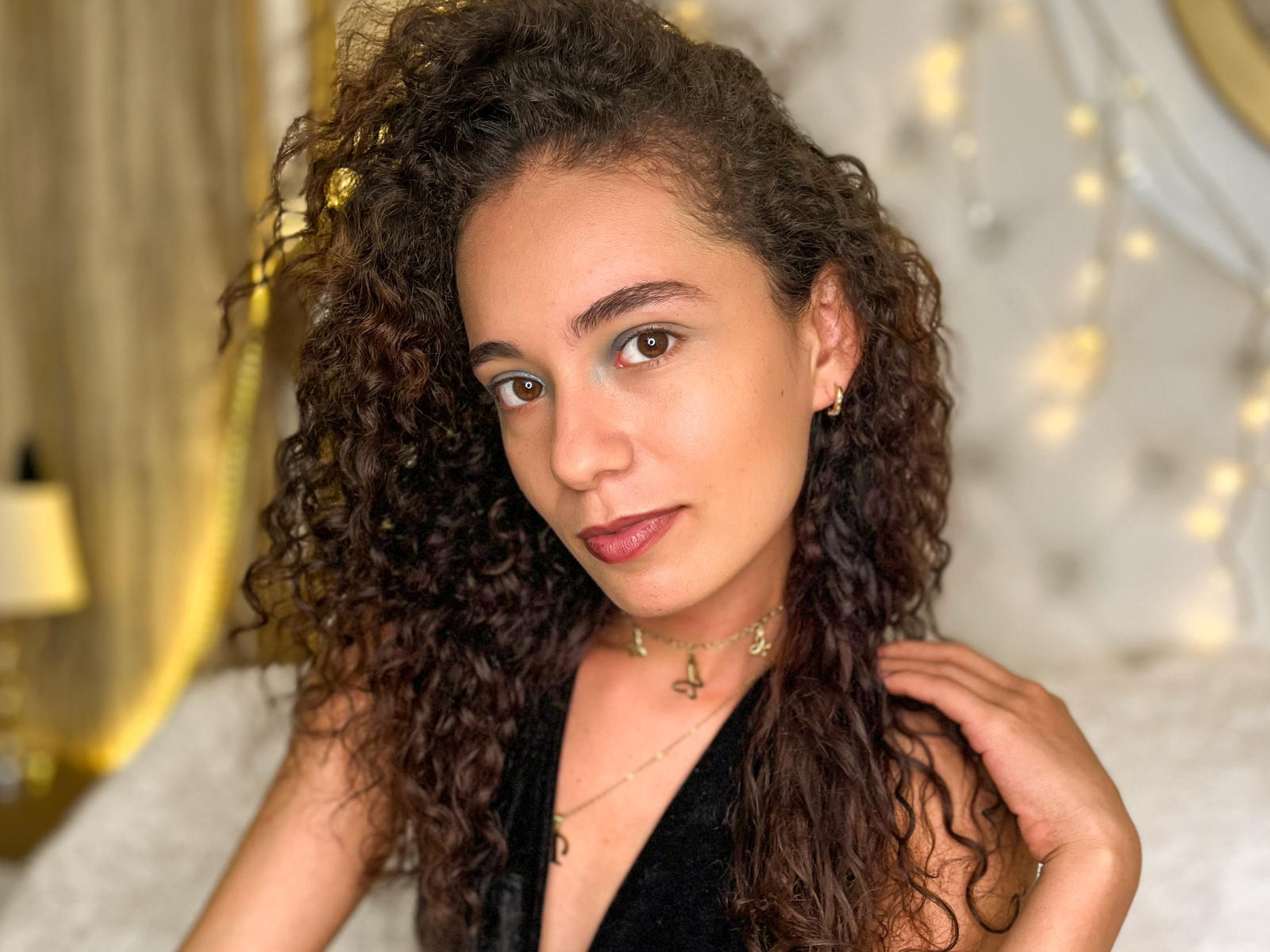 AlayaBraw, de cabello rizado, con piel morena, ojos marrones y lápiz labial rojo, vestida con una camiseta negra sin mangas, un collar dorado y aretes, mirando a la cámara con una ligera sonrisa..