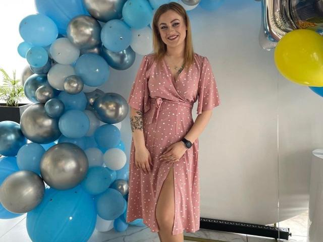 Swoiia with light brown hair, wearing a pink polka dot dress with a thigh slit, smiling, holding her dress, blue and silver balloons, tattoo on left arm.