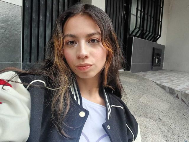 Katherin Johns, con cabello castaño largo, vestida con una chaqueta deportiva negra y blanca, tiene un rostro adorable, piel clara y un maquillaje sutil, y está de pie en un entorno exterior gris y texturizado..