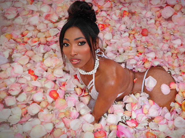 SabrinaDevil, à la peau foncée, recouverte de pétales de rose rose, portant un bikini blanc et un collier de perles, regardant vers le haut avec un sourire séduisant, ses fesses relevées, dans un lit rempli de pétales..