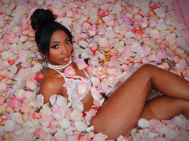 SabrinaDevil lying in pink and white rose petals, wearing white lace lingerie and a pearl necklace, with dark skin, brown eyes, and black hair in a bun, looking at camera.