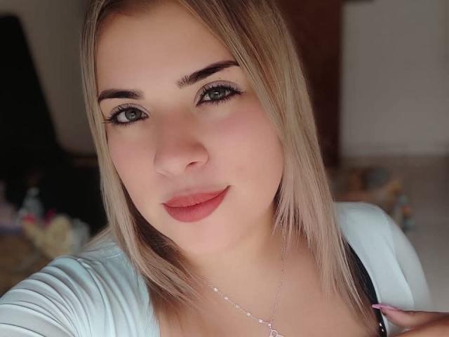 TatianaTee heeft blond haar, groene ogen en roze lippen. Ze draagt een lichtblauwe top, waar een ketting zichtbaar is. Haar borsten zijn klein en ze glimlacht zachtjes..