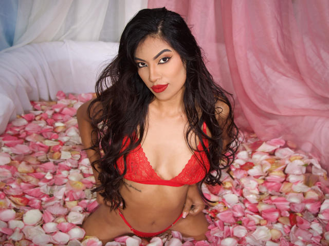 KendallJones with long black hair, red lace bra, and matching thong, sitting in pink and white rose petals, red lips, and a confident, sexy look, wearing red lipstick.
