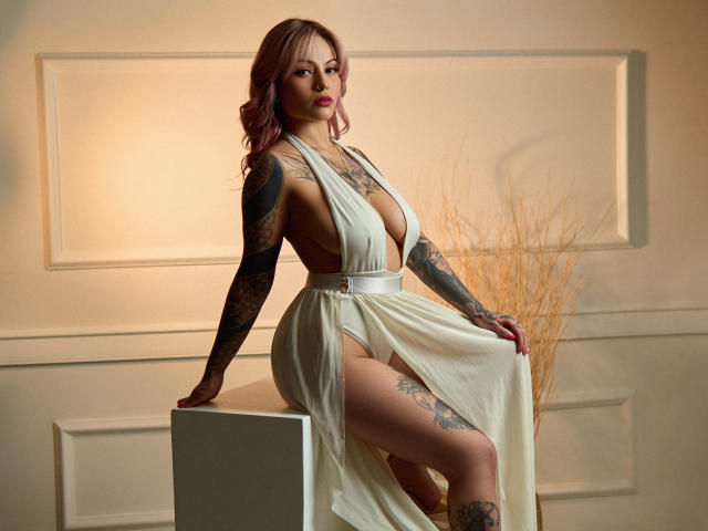 KozukiWen, with pink hair, tattoos on arms and legs, wearing a deep-cut, white, side-slit dress, sitting on a white cube, exposing her tits and a glimpse of her pussy.